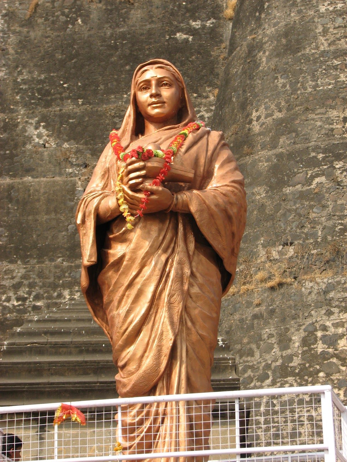 Garlanded bronze statue of Ahilyabai Holkar before a stone fort wall.